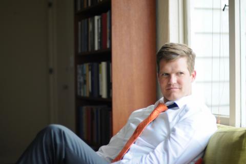 Peter Wayne Moe a white male sitting on a green couch in front of a windoe next to a brown book case with a white button down with an orange tie and gray slacks looking away with a smirk