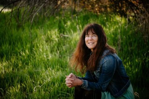 photo of mary emerick sitting in green grass. She has long brown hair with bangs and a jean jacket on