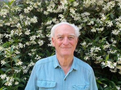 Head shot photo of author Stephen Most. He is wearing a blue collared shirt in front of green shrubbery with white flowers.